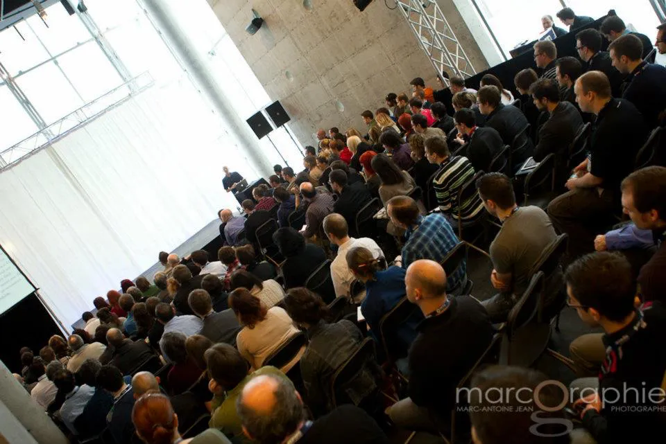 A packed auditorium audience seated in rows, watching a speaker on a lit stage.