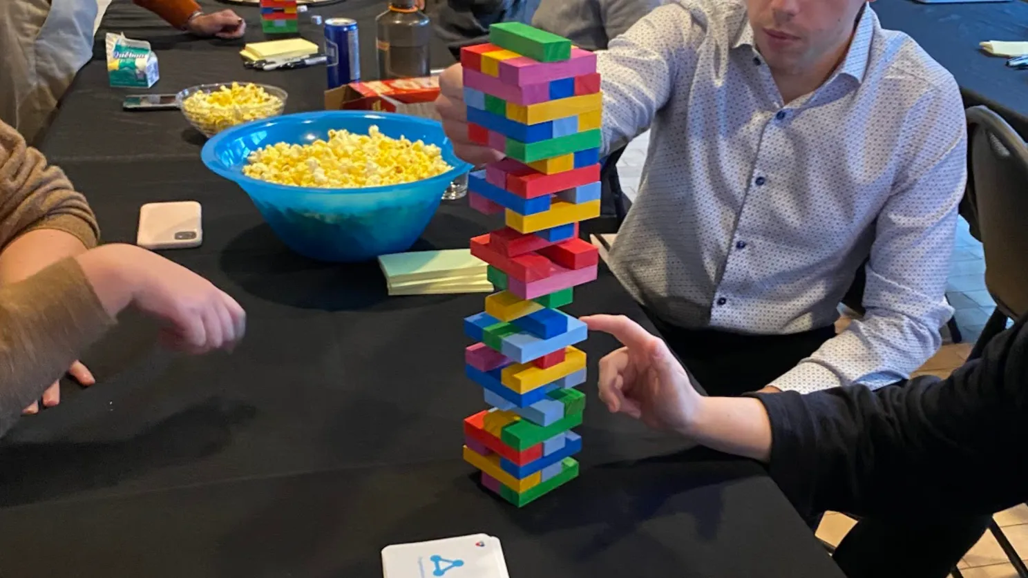Players reaching for a tall, colorful Jenga tower on a table with a bowl of popcorn.