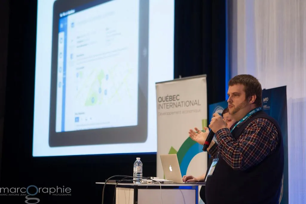 A man speaking into a microphone beside a laptop and a “Québec International” banner, with a slide showing a mobile map interface projected behind him.