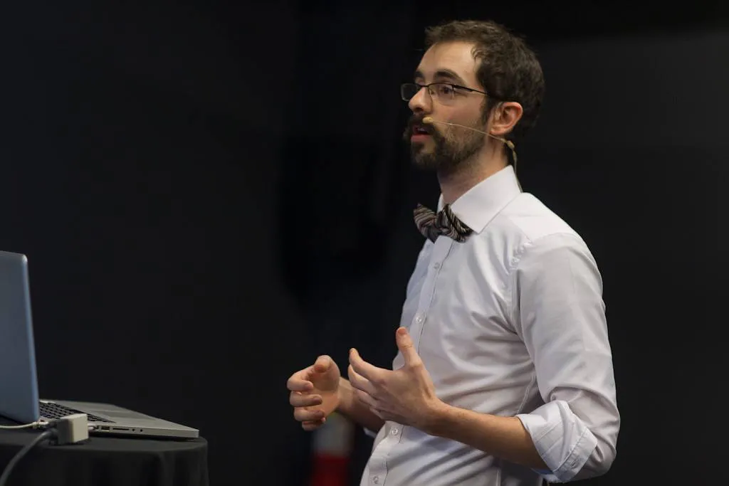 A bearded man in a bow tie and headset microphone speaking with his hands raised mid-gesture.