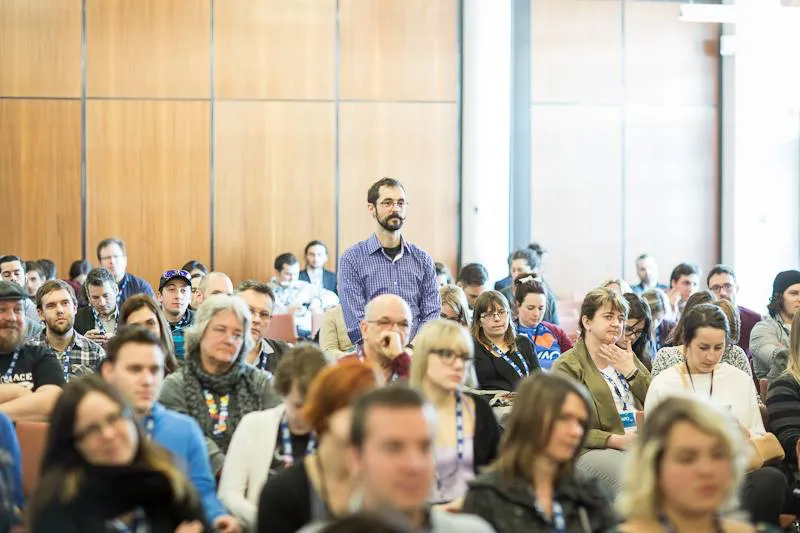 An audience member standing up to ask a question in a wood-paneled conference hall.