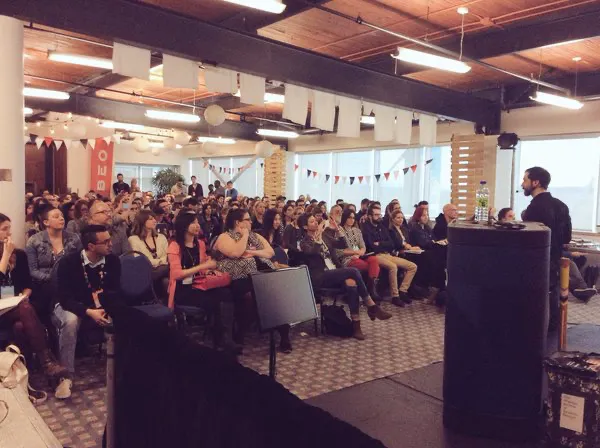 A speaker at a podium addressing a seated audience in a room decorated with pennant flags.