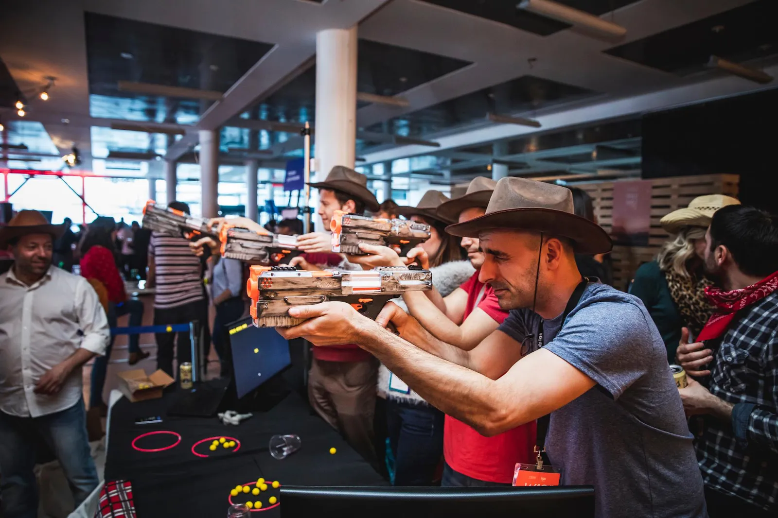 A row of people in cowboy hats aiming toy blaster guns at a target on a table.