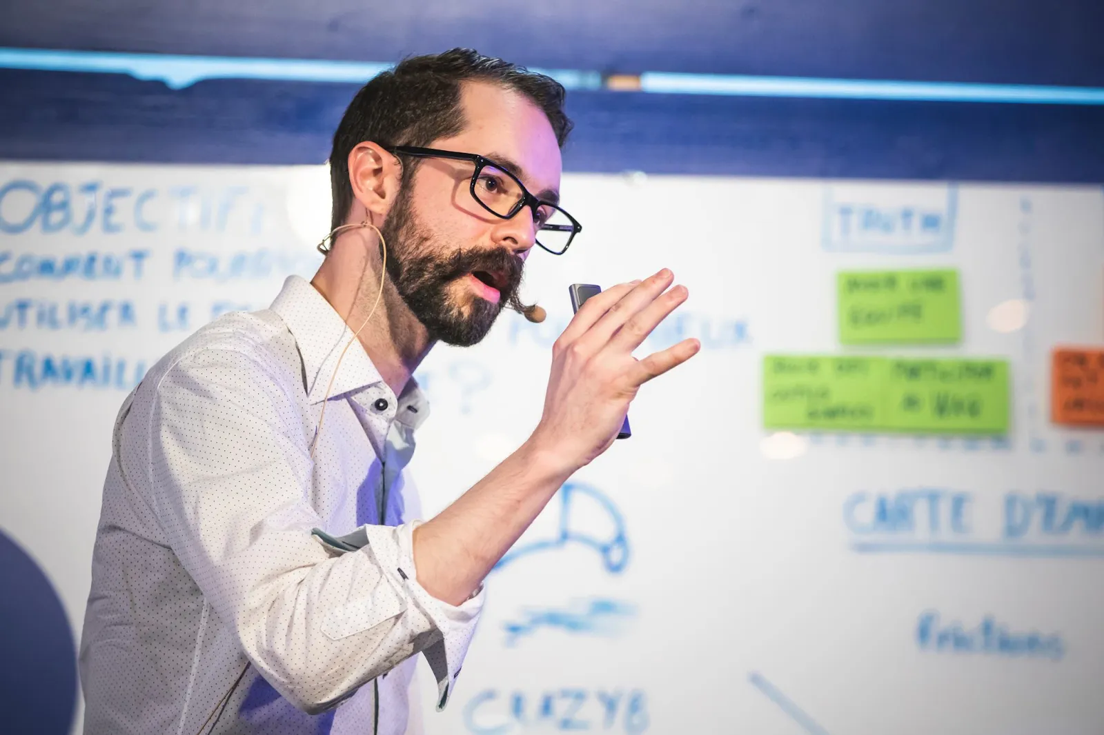 A man with a headset microphone gesturing while speaking in front of a whiteboard covered in sticky notes and handwritten headings like “OBJECTIF” and “CARTE D’EMPATHIE.”