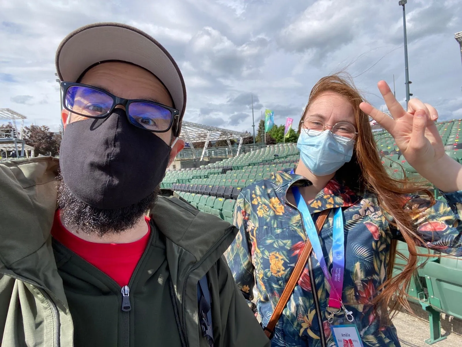 Two masked people taking a selfie in empty stadium bleachers, one flashing a peace sign.