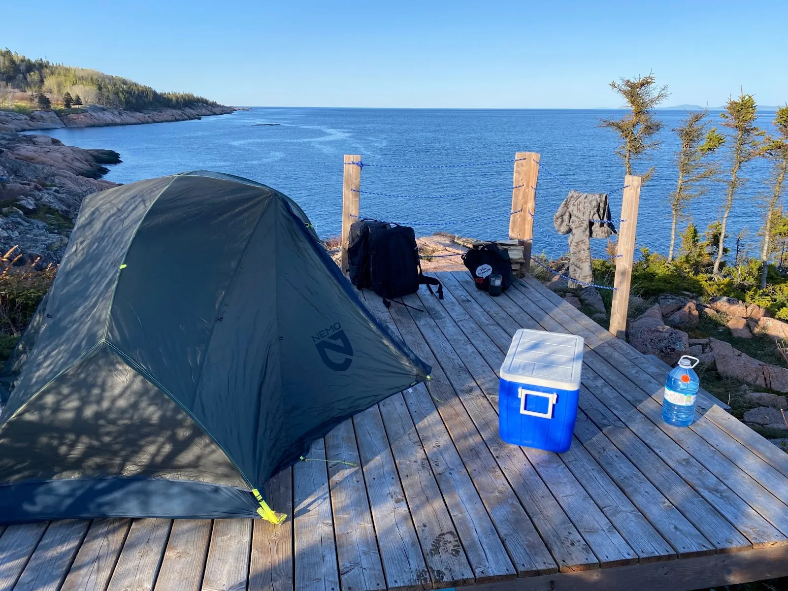 A camping tent pitched on a wooden dock overlooking the ocean, with backpacks and a cooler nearby.