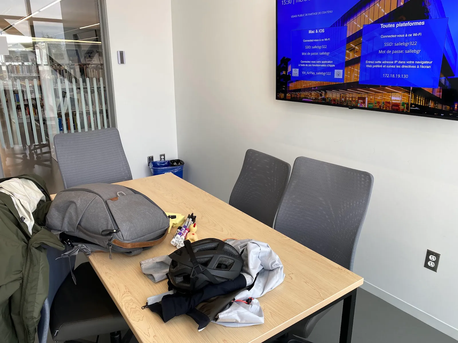 A meeting room table with a backpack, a bike helmet, and a jacket, with a wall screen showing Wi-Fi connection instructions.