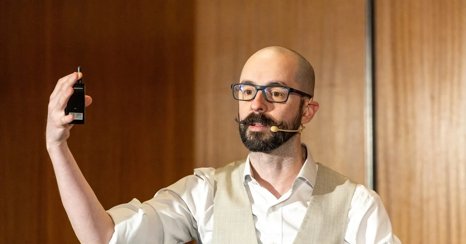 A bald, bearded man in a vest holding up a small black device while speaking with a headset microphone.