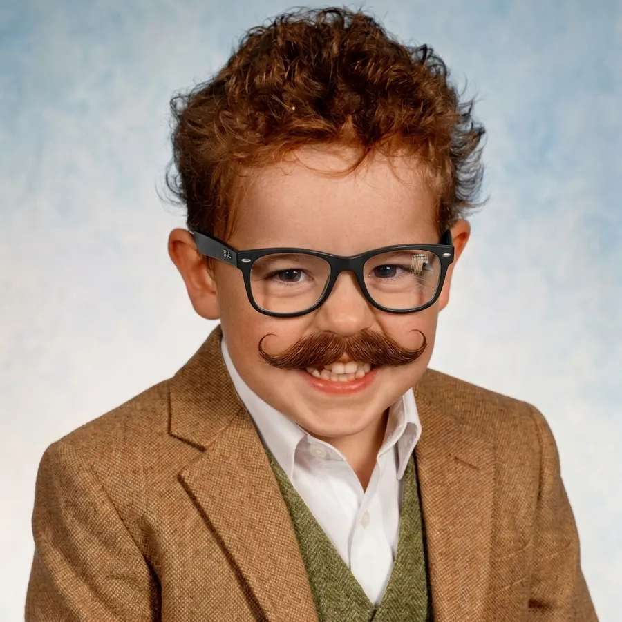 A young boy in glasses and a fake moustache, dressed in a matching blazer and vest